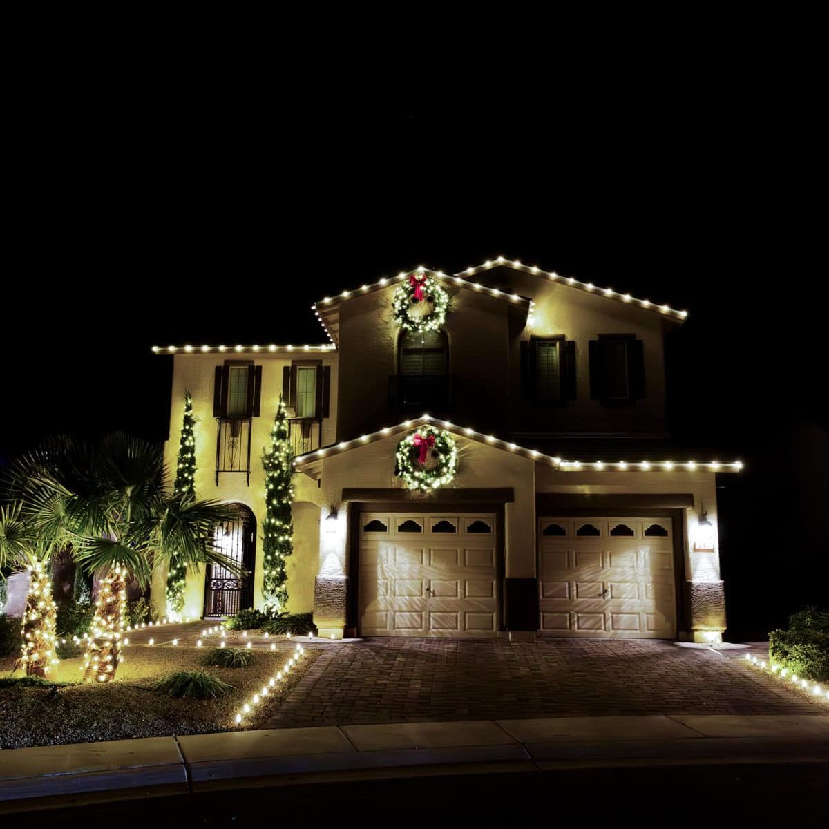 Warm white lights installed with matching lit christmas wreaths installed above garage and top of house. Warm white christmas lights installed on palm trees and along driveway